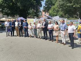 Słoneczny dzień, na przemyskim rynku grupa osób stoi w szeregu, za mini wystawa prac