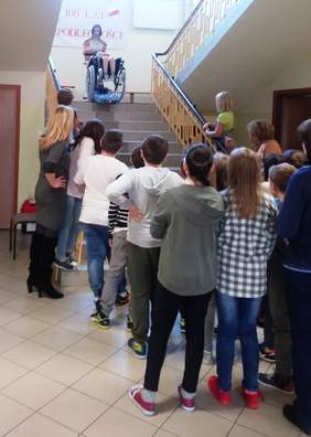 Pokaż zdjęcie: Grupa osób stojąca przy schodach, na górze schodów znajduje się osoba na wózku zjeżdzająca na schodołazie