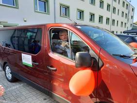 Pokaż zdjęcie: W wiśniowym samochodzie od strony pasażera siedzi pan, który życzliwie sie uśmiecha. 