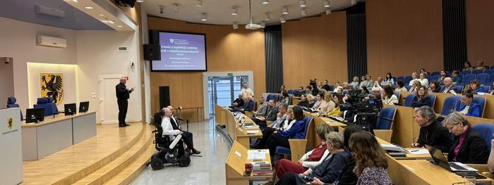 Pokaż zdjęcie: Sala konferencyjna z dużą liczbą osób. Na środku widać osobę na wózku, a na ekranie wyświetlony jest temat dotyczący osób z niepełnosprawnościami. Pomieszczenie jest przestronne i nowoczesne.