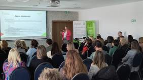 Pokaż zdjęcie: Widok na sale od tyłu, widać gości siedzących na krzesłach i w tle ekran z prezentacją