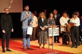 Pokaż zdjęcie: Na scenie stoją elegancko ubrani ludzie. Jeden z mężczyzn trzyma w dłoni mikrofon, do którego przemawia. Jedna osoba siedzi na wózku inwalidzkim. 