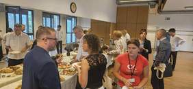 Pokaż zdjęcie: Na stołach znajduje sie poczęstunek dla uczestników Targów Pracy i Możliwości dla Osób z Niepełnosprawnościami. Wokół zebrali się ludzie, którzy jedzą i rozmawiają ze sobą. 