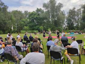 Pokaż zdjęcie: Duże boisko otoczone wysokimi drzewami na pierwszym planie publiczność siedzi w kilku rzędach na krzesłach odwrócona plecami przed nimi stoją cztery kobiety za nimi jest różowa mata  i aktorzy siedzący na matach