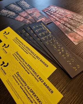 Pokaż zdjęcie: Na stole leżą prostokątne, żółte zakładki z czarnym napisem zapraszamy na spektakle bez barier, napis jest również zapisany w języku braille'a obok leżą czarne zakładki.