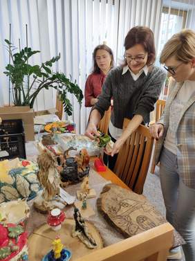 Pokaż zdjęcie: Trzy kobiety oglądają i oceniają prace plastyczne wykonane przez uczestników konkursu — na stole znajdują się kolorowe rzeźby, ceramika i płaskorzeźby.