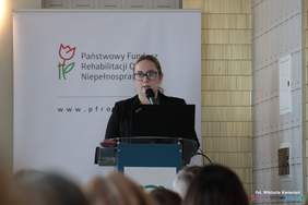 Pokaż zdjęcie: Przy mównicy stoi kobieta uczesana na gładko w okularach