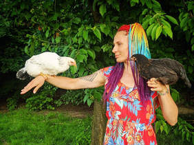Pokaż zdjęcie: Zdjęcie na łonie natury, kobieta z kolorowymi dredami i kolorowej sukience patrzy w lewą stronę, w stronę ptaka, który siedzi jej na prawej ręce, w lewej dłoni trzyma drugiego ptaka