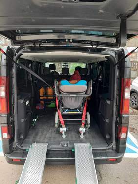 Pokaż zdjęcie: Na parkingu stoi samochód dostosowany do potrzeb pasażera z niepełnosprawnością. Tylna klapa jest otwarta i we wnętrzu pojazdu siedzi osoba na wózku inwalidzkim. Do auta dostawione są szyny, dzięki którym można wprowadzić wózek.  