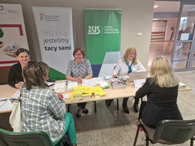 Na tle banerów organizatorów wydarzenia siedzą za stołami trzy usmiechnięte kobiety, przed nimi siedzą dwie inne kobiety, widzimy je z tyłu