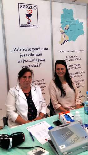 Pokaż zdjęcie: Punkt pomiaru ciśnienia – dwie pielęgniarki na tle baneru reklamowego Samodzielnego Zespołu Publicznych Zakładów Lecznictwa Otwartego Warszawa-Ochota