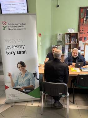 Pokaż zdjęcie: Trzech mężczyzn znajduje się przy stoliku z materiałami informacyjnymi. Jeden siedzi tyłem do aparatu, a dwóch pozostałych siedzi po drugiej stronie stołu. Obok widoczny jest baner.