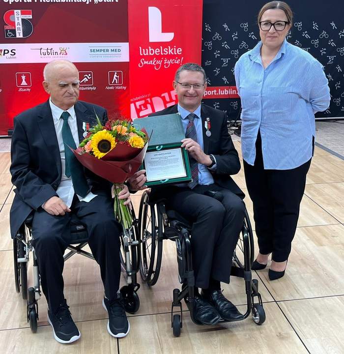 Pokaż zdjęcie: Dwóch mężczyzn na wózkach trzyma w rękach kwiaty i grawerton, obok stoi kobieta w niebieskiej koszuli.