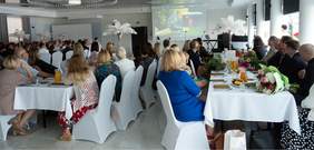 Pokaż zdjęcie: Grupa uczestników spotkania siedzi w sali bankietowej na białych krzesłach. Oglądają film, dokumentujący działalność WTZ w Augustowie.