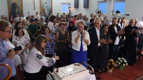 Pokaż zdjęcie: Grupa ludzi zgromadzona w sali,  podczas uroczystości jubileuszowej. Na pierwszym planie kobieta w haftowanej bluzce kroi duży prostokątny tort z napisem "25 lat ŚDS i WTZ w Konarach". Wokół niej stoją i klaszczą inni uczestnicy wydarzenia – kobiety, mężczyźni, niektórzy w eleganckich strojach, inni w habitach zakonnych. W tle widać tłum ludzi oraz obrazy religijne zawieszone na ścianach. Atmosfera jest uroczysta i radosna.