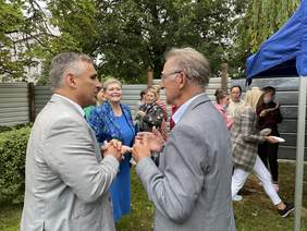 Pokaż zdjęcie: Dwóch mężczyzn w ubranych w szare garnitury oraz kobieta w granatową wzorzystą garsonkę rozmawiają ze sobą. 