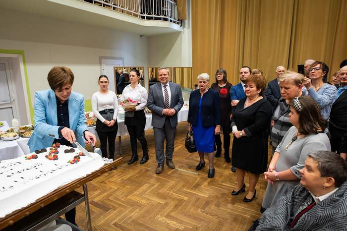 Pokaż zdjęcie: Grupa siedemnastu osób w różnym wieku stoi w sali, w której zorganizowany został poczęstunek. Jedna z kobiet ubrana w niebieską marynarkę trzyma w dłoniach nóż, którym kroi okolicznościowy tort na mniejsze kawałki. W tle widoczny jest balkon oraz jasnopomarańczowe zasłony sięgające podłogi pokrytej brązową wykładziną o wzorze przypominającym schody.