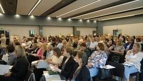 Pokaż zdjęcie: W auli ustawione są rzędy krzeseł, na których siedzą ludzie. Niektórzy robią notatki, a inni wykonują zdjęcia. 