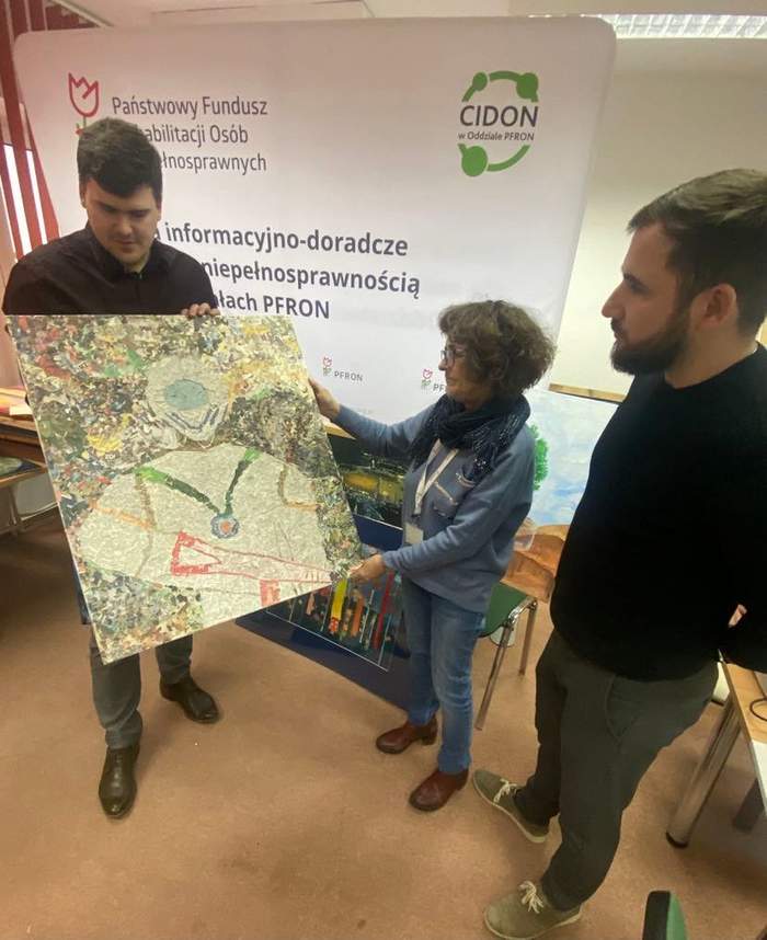 Pokaż zdjęcie: Kobieta i mężczyzna trzymają w ręku obraz, podziwiają go, po prawej mężczyzna, który patrzy na pracę, za nimi baner CIDON