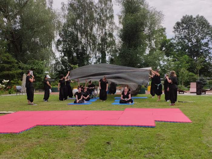 Pokaż zdjęcie: Na trawie rozłożona jest różowa mata za matą na tle dużych zielonych drzew są aktorzy ubrani w czarne stroje część aktorów siedzi na niebieskich matach pozostali stoją i naciągają na nich czarną folię 