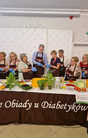 Pokaż zdjęcie: Grupa uczestników wydarzenia „Czwartkowe Obiady u Diabetyków” stoi przy stole zastawionym warzywami i ziołami, przygotowując potrawy w ramach warsztatów kulinarnych.