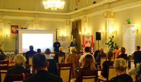 uczestnicy uroczystości, siedzą w sali, żółte oświetlenie, mężczyzna przemawia