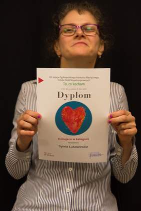 Pokaż zdjęcie: Uśmiechnięta kobieta w dłoniach przed sobą trzyma dyplom.