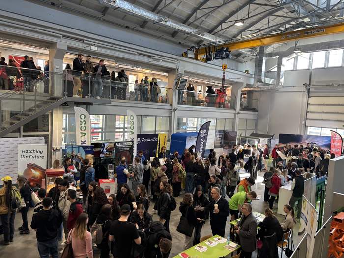 Pokaż zdjęcie: Widok z góry na uczestników Targów Pracy na Uniwersytecie Przyrodniczym w Poznaniu. W hali widać tłum osób rozmawiających przy stoiskach wystawienniczych, flagi oraz roll-upy promocyjne.