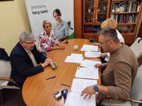 Pokaż zdjęcie: Przy owalnym stole siedzą dwie kobiety i dwóch mężczyzn. Na stole leżą dokumenty, pieczątki, długopisy, telefony. Na regale stoją książki, grawery, medale, odznaczenia. 