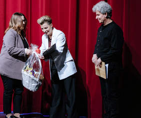 Pokaż zdjęcie: Trzy osoby na scenie, w tle czerwona kotara. Dwie kobiety przekazują sobie kosz ze słodyczami.