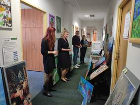 Pokaż zdjęcie: W długim korytarzu, po jego lewej i prawej stronie na podłodze i na krzesłach poustawiane są prace plastyczne. Pośród prac spaceruje komisja, która ocenia prace w poszczególnych kategoriach. Na zdjęciu widać cztery osoby.