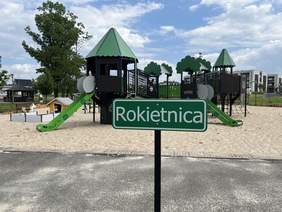 Pokaż zdjęcie: Na nawierzchni z piasku znajdują się zestawy zabawowe takie jak: domek na drzewie, kawiarenka i remiza strażacka wraz ze zjeżdżalniami. Z przodu widoczna jest tabliczka z napisem Rokietnica. W tle znajdują się drzewa oraz budynki mieszkalne.