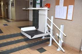 Pokaż zdjęcie: Urządzenie pozwalające na transport osoby z niepełnosprawnością ruchu, z pominięciem przeszkody w formie trzech stopni różnicujących poziomy pomiędzy dwoma pomieszczeniami. W tle tablica korkowa z zapisanymi kartkami oraz drzwi do innych pomieszczeń.