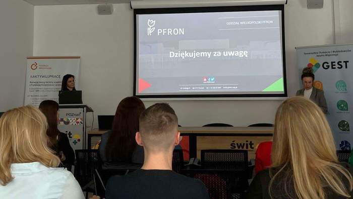 Pokaż zdjęcie: Dwie kobiety stoją po obu stronach dużego ekranu, na którym wyświetlone jest: logo PFRON, nazwa Oddział Wielkopolski PFRON oraz sformułowanie „Dziękujemy za uwagę”. Na sali widoczna jest grupa sześciu osób, która przysłuchuje się wystąpieniu.