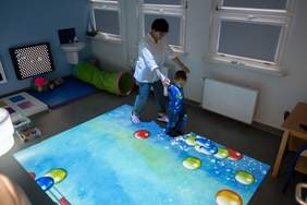 Pokaż zdjęcie: Kobieta prowadząca małego chłopca po podświetlanej macie z różnokolorowymi balonami oraz przewagą koloru błękitnego. W tle materac, umywalka oraz małe okna przysłonięte żaluzjami.