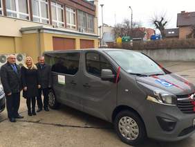 Pokaż zdjęcie: Szary bus, obok stoi kobieta i dwówch mężczyzn.