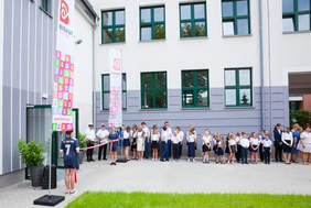 Pokaż zdjęcie: Uczniowie ustawieni są wzdłuż nowego budynku, przed samym wejściem stoją dwie uczennice i trzymają białoczerwoną wstęgę