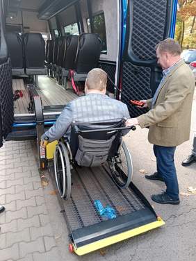 Pokaż zdjęcie: Mężczyzna na wózku inwalidzkim wjeżdża za pomocą windy do wnętrza samochodu. Obok stoi pan, który za pomocą pilota steruje urządzeniem.  