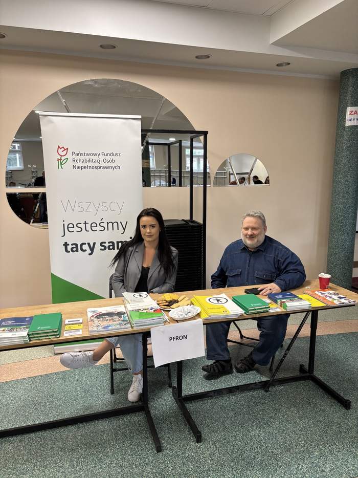 Pokaż zdjęcie: Dwie osoby siedzą przy stole z rozłożonymi broszurami, ulotkami i publikacjami. Za nimi stoi roll-up z logo PFRON i napisem „Wszyscy jesteśmy tacy sami”. Zdjęcie wykonane wewnątrz budynku.