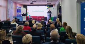 Pokaż zdjęcie: Zdjęcie przedstawia konferencję ZUS w Białymstoku. Przy mównicy przemawia Marek Marczewski, Dyrektor Oddziału ZUS. W tle ekran z napisem „OTWARCIE KONFERENCJI” i banery TVP3 Białystok oraz ZUS. Na sali siedzą elegancko ubrani uczestnicy, a po lewej stronie ekipa telewizyjna i monitor z tłumaczem języka migowego.