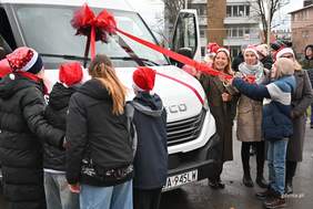 Pokaż zdjęcie: Grupa osób w czapkach świątecznych stoi przy przodzie dużego białego pojazdu udekorowanego czerwonymi wstążkami i dużą kokardą na szybie. Kilka osób wspólnie trzyma czerwoną wstęgę, uśmiechając się podczas uroczystości.