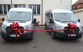 Pokaż zdjęcie: Na zdjęciu dwa srebrne busy zaparkowane obok siebie, połączone długą czerwoną wstęgą z dużymi czerwonymi kokardami na przodach. Wstęga biegnie poziomo między pojazdami. Busy stoją na tle jasnożółtego budynku.