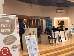 Pokaż zdjęcie: Za mównicą stoi mężczyzna w koszuli i marynarce, mówi do mikrofonu. Po lewej stronie baner promujący wydarzenie po prawej baner Radia Gdańsk i godło Województwa Pomorskiego, nad nim wisi krzyż.