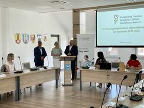 Pokaż zdjęcie: Stoją trzy osoby. Mężczyźni ubrani w marynarkach, w środku kobieta w jasnej bluzce okularach. Za nimi wiszą herby. Po prawej telebim na którym wyświetlane jest logo PFRON. Przed nimi stoi mównica, po prawej i lewej stronie stoją biurka za który