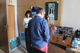 Pokaż zdjęcie: Zdjęcie pokoju biurowego, na krzesłach i podłodze poustawiane są prace plastyczne, komisja plastyczna przygląda się pracom