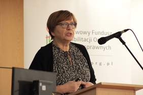 Pokaż zdjęcie: Przy mównicy kobieta w średnim wieku w okularach