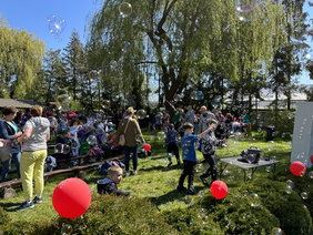 Pokaż zdjęcie: Polana dookoła dużo wysokich drzew pośrodku niej ławki siedzący na niej ludzie. Dzieci biegają i łapią bańki mydlane, które wylatują z automatu.