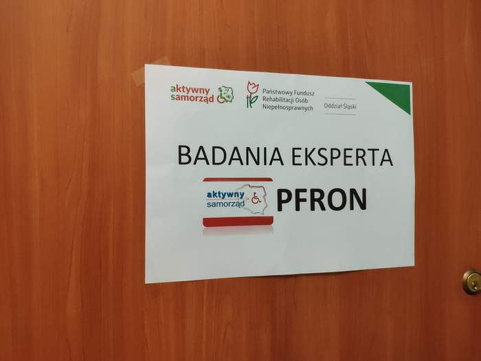 Pokaż zdjęcie: na fragmencie brązowych drzwi zawieszony jest plakat z czarnym napisem badania eksperta PFRON 