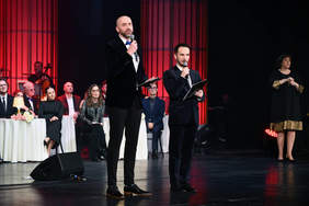 Dwóch mężczyzn na scenie, w czarnych garniturach, w tle stoliki, przy których siedzą kobiety i mężczyźni