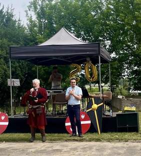 Pokaż zdjęcie: Dwóch mężczyzn stoi przed sceną plenerową z balonami w kształcie liczby 20. Jeden z nich ubrany jest w historyczny strój, trzyma mikrofon i przemawia. Drugi klaszcze, stojąc obok. Za nimi widoczna scena z zadaszeniem, elementy dekoracyjne w postaci tarcz.